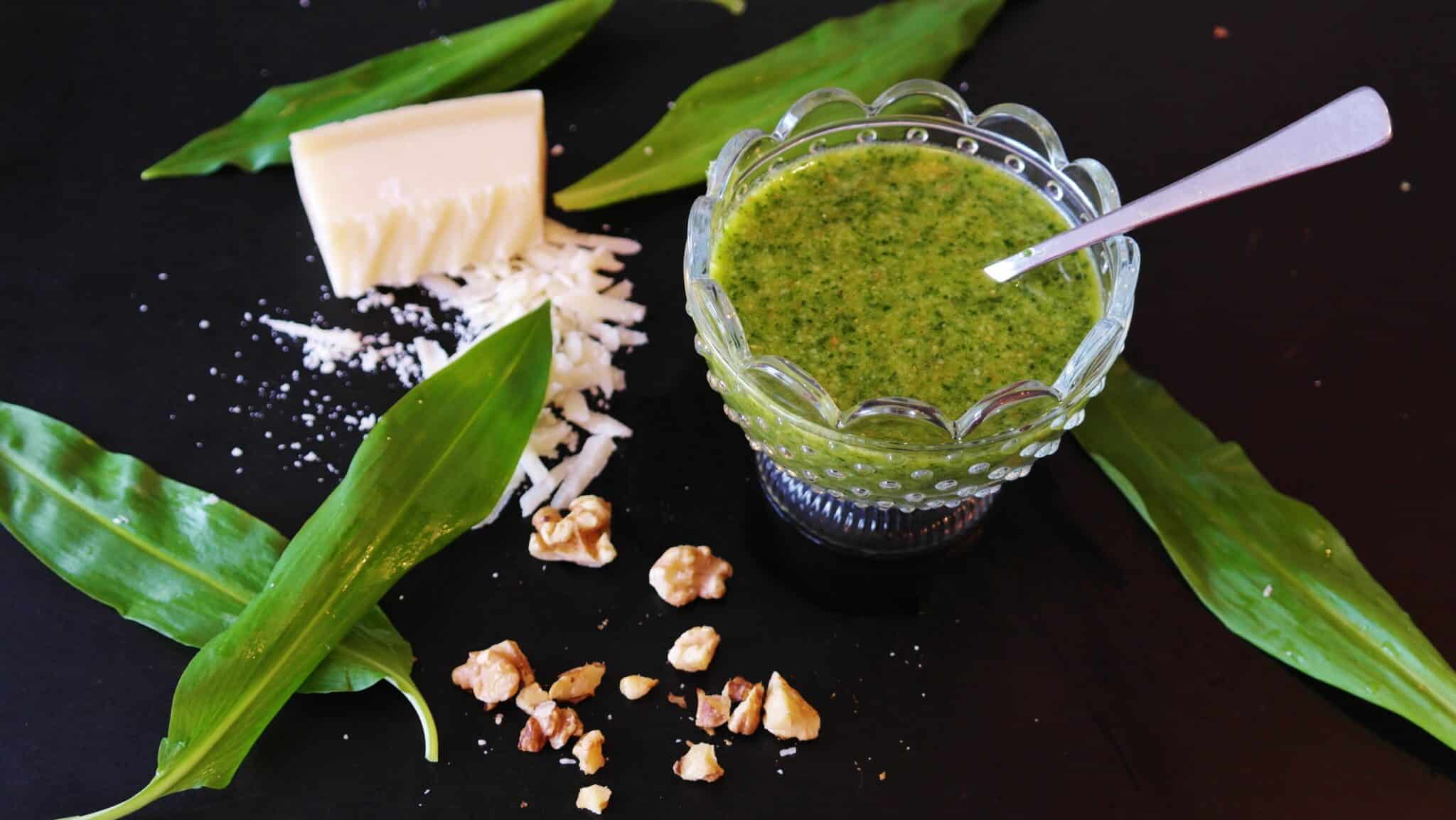 Grünes Pesto in einem Glasschälchen, daneben Bärlauchblätter, ein Stück Parmesan und Walnüsse